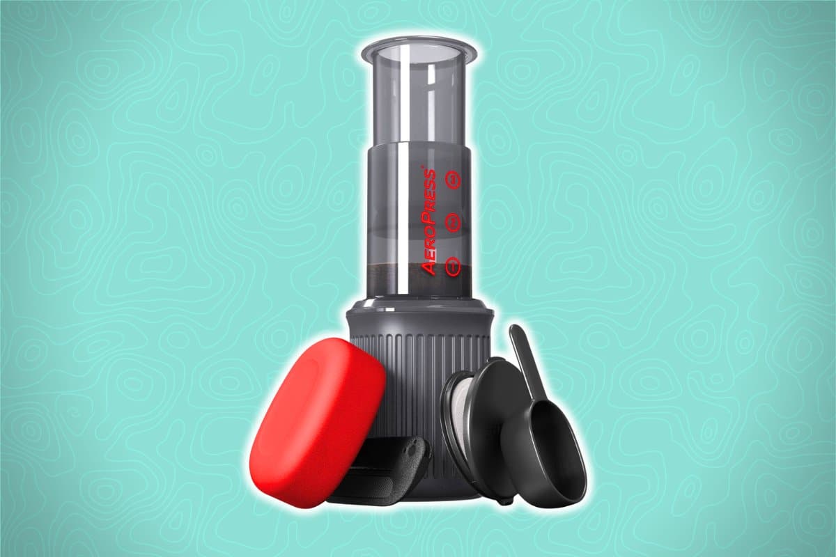 Aeropress Go coffee maker with accessories, featuring a red travel case, black scoop, and black stirrer, set against a light teal background with a subtle wavy pattern.