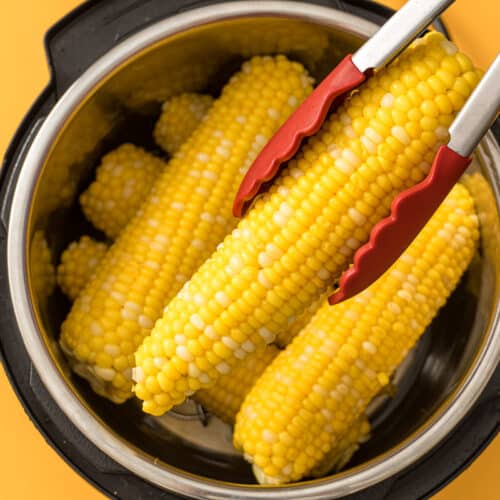 A pot filled with freshly cooked corn on the cob, with yellow and white kernels visible, and a pair of tongs with red tips lifting one cob out of the pot against a yellow background.