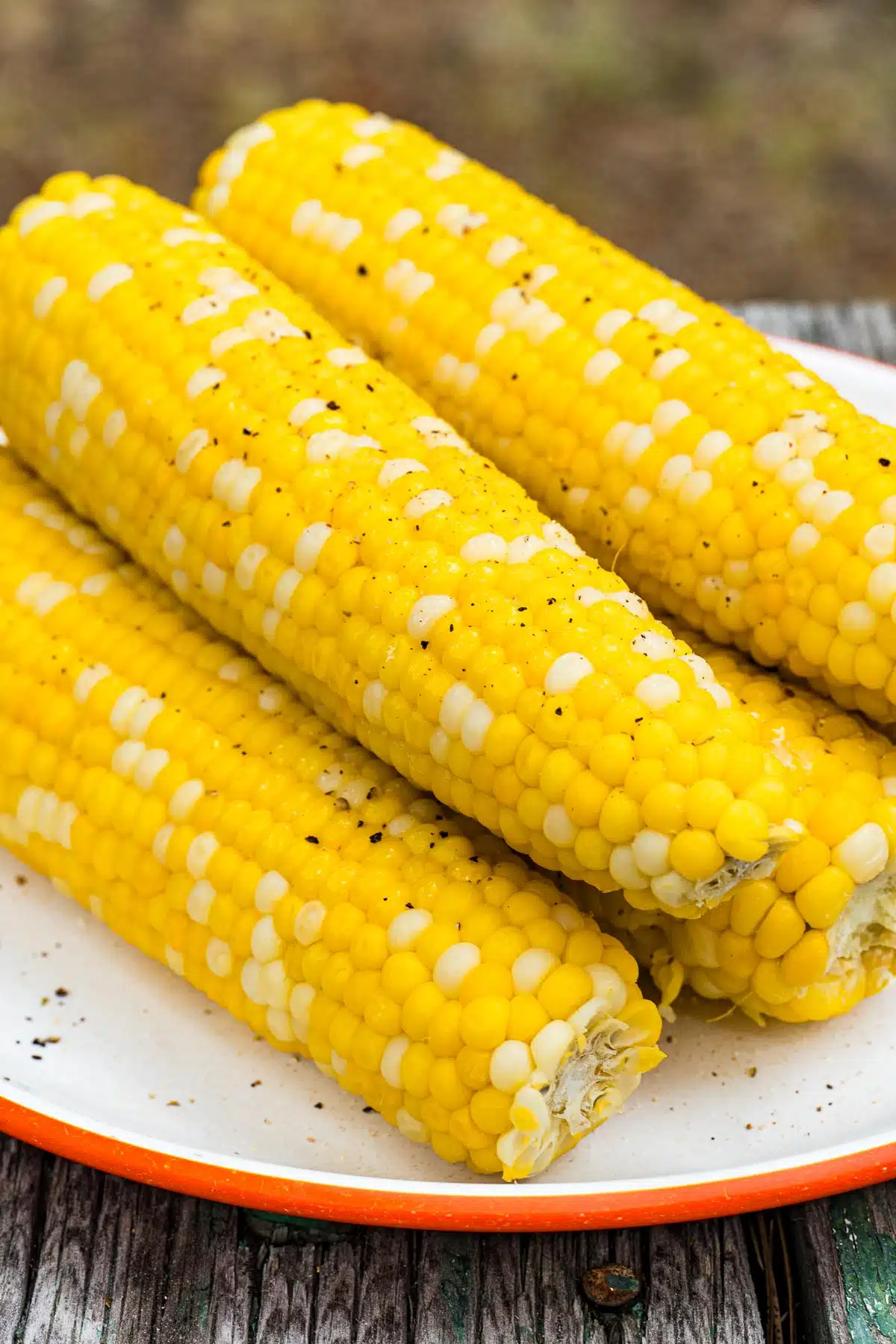 Four ears of corn on the cob, featuring bright yellow and white kernels, are placed on a white plate with an orange rim, set on a wooden surface.