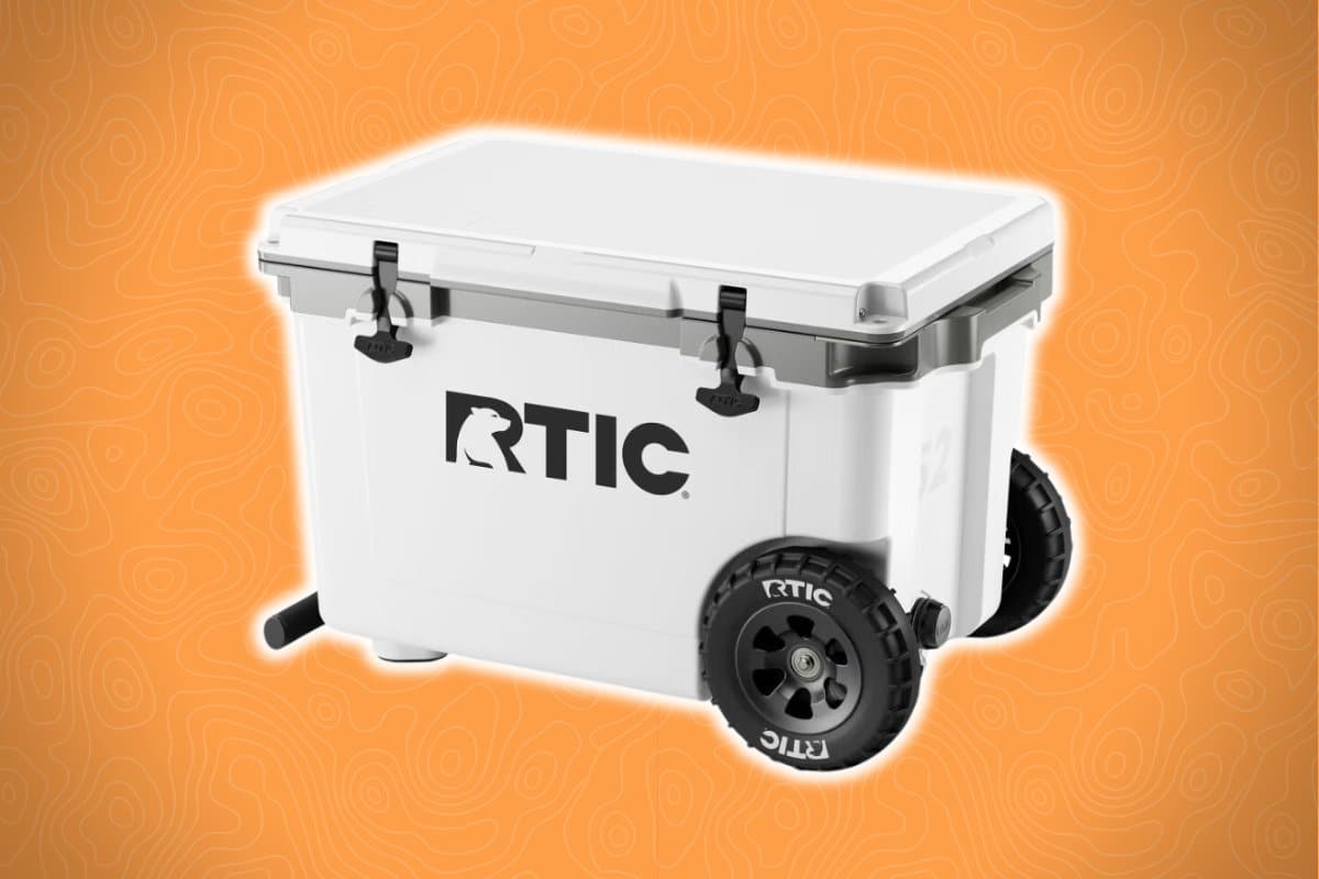 A white RTIC cooler with black wheels and two black latches on the lid sits against an orange background with a subtle abstract pattern.