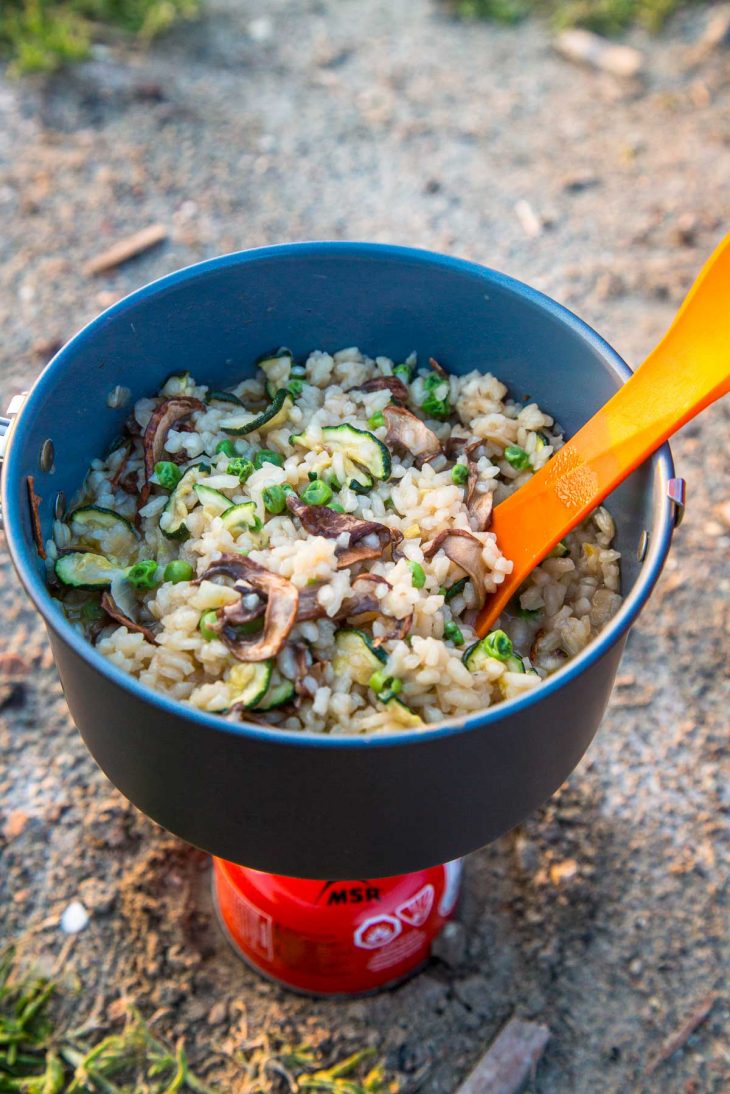 A bowl of chili topped with shredded cheese, diced avocado, sour cream, corn chips, and chopped green onions sits on a rustic wooden surface with a lime wedge on the side and a spoon tucked into the bowl.