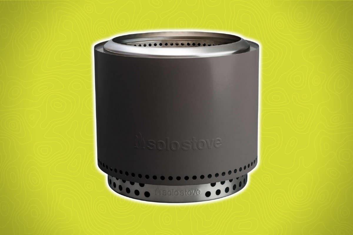 A sleek, dark gray Solo Stove with a cylindrical design and ventilation holes around the top and bottom edges sits against a light green background.