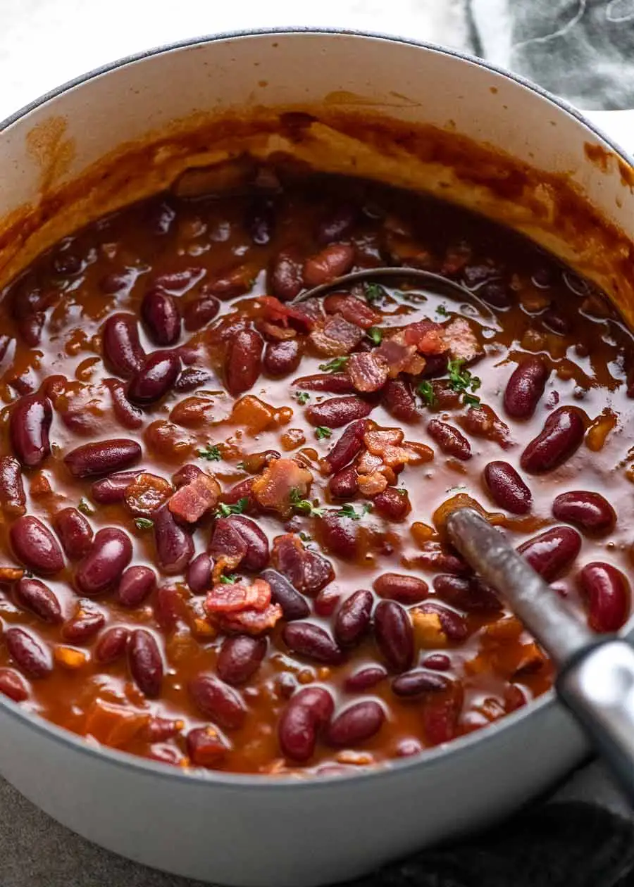 A pot contains kidney beans in a thick sauce, some slightly crushed, topped with visible bacon pieces and pierced by a dipped ladle.
